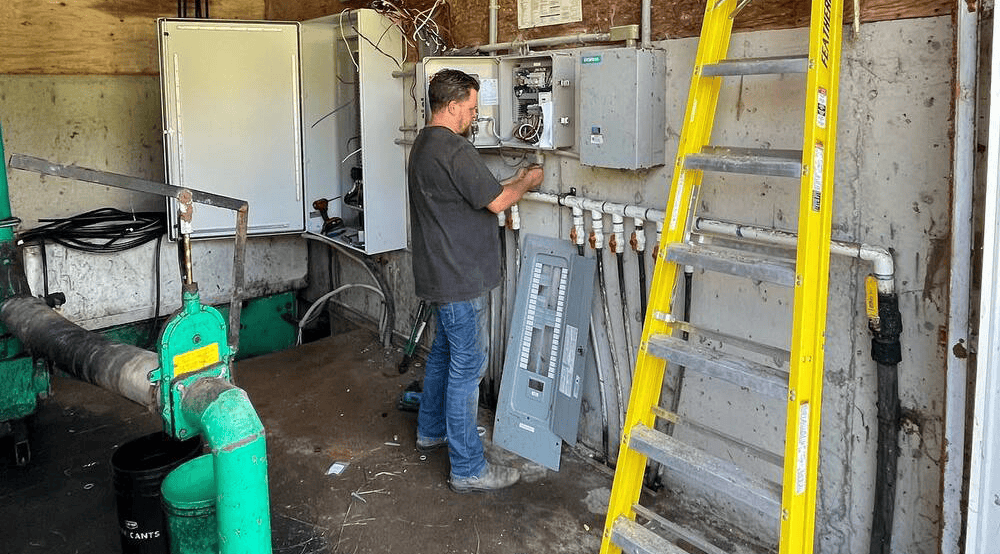 An electrician working in a utility room, inspecting electrical panels and equipment, with tools and a ladder nearby.